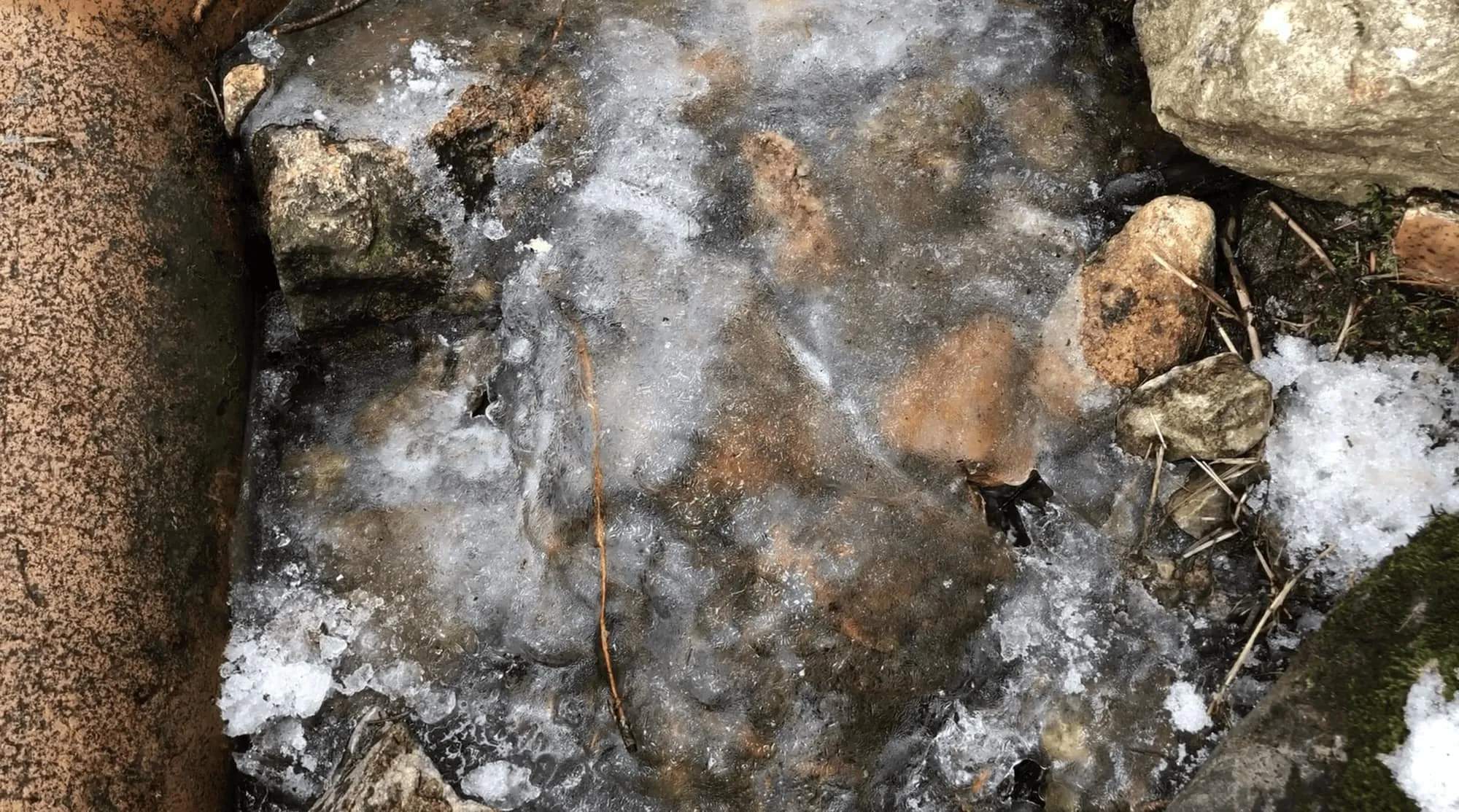 Close-up view of a small frozen stream with visible rocks and pebbles beneath the ice. Surrounding the stream are various-sized stones and remnants of snow.